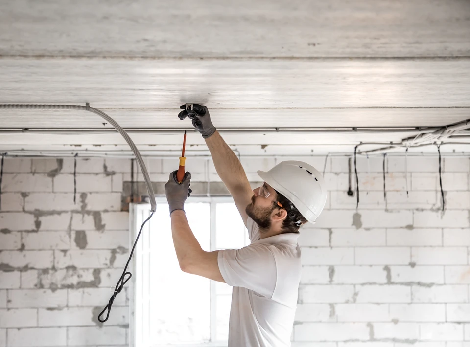 Électricien professionnel en casque blanc installant un système électrique au plafond d'un bâtiment en construction, murs en parpaings apparents, câblage apparent