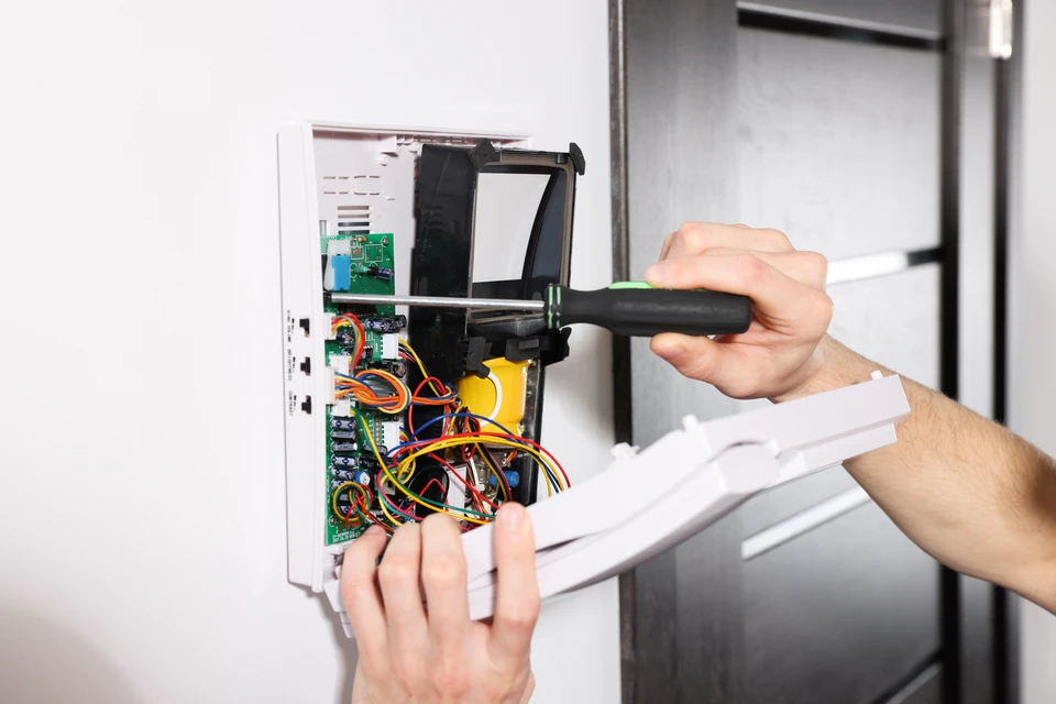Gros plan d'un technicien électricien en tenue professionnelle tirant un câble de sécurité blindé à travers une gaine murale grise de type goulotte, dans un couloir technique tertiaire avec faux plafond modulaire et équipements muraux professionnels en arrière-plan.