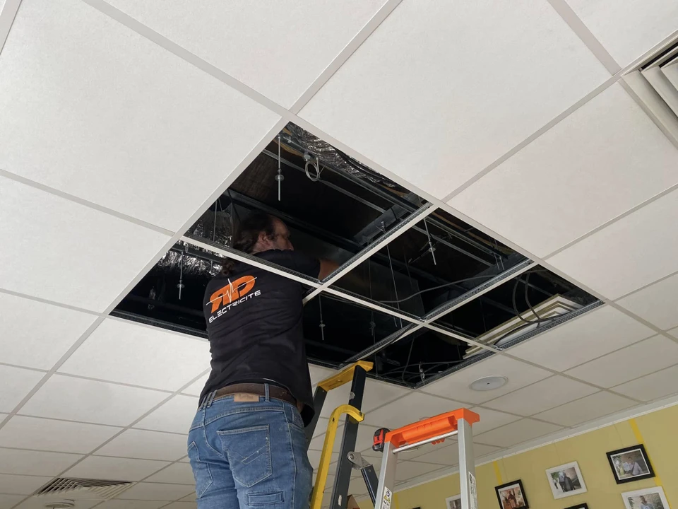 Technicien en vêtements noirs sur escabeau orange installant une unité de climatisation cassette encastrée dans un faux plafond dalle